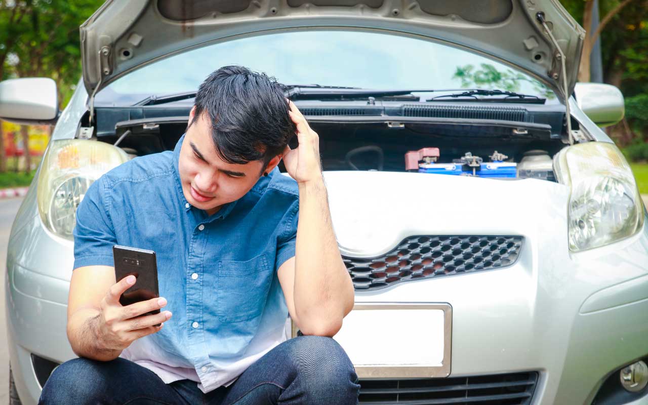 A young man, dressed in a blue shirt, sits in front of a car with the hood open, revealing the engine compartment. He looks concerned while looking at his smartphone, possibly searching for a solution to a car problem or calling for assistance.