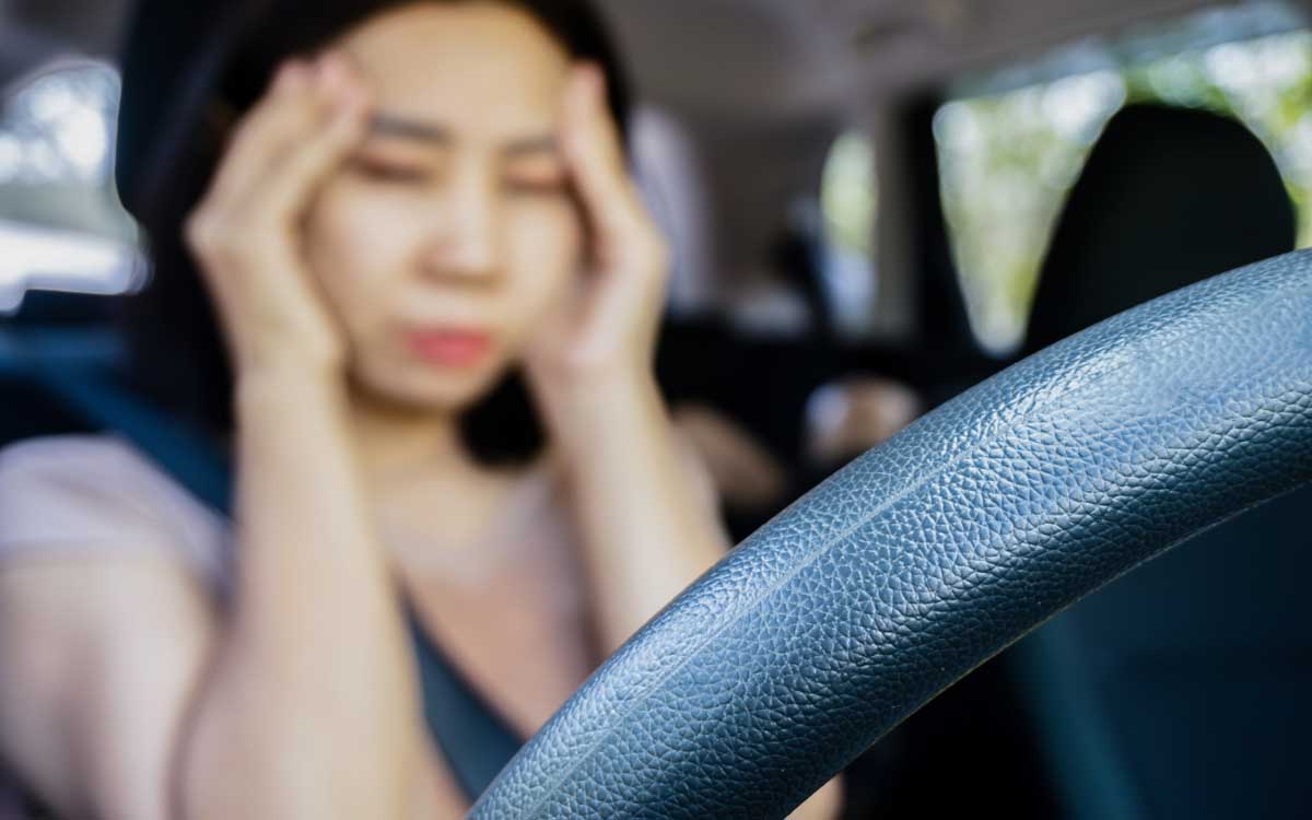 A woman sitting in the driver's seat of a car, holding her head with both hands, eyes closed, and appears to be experiencing a headache or tired.