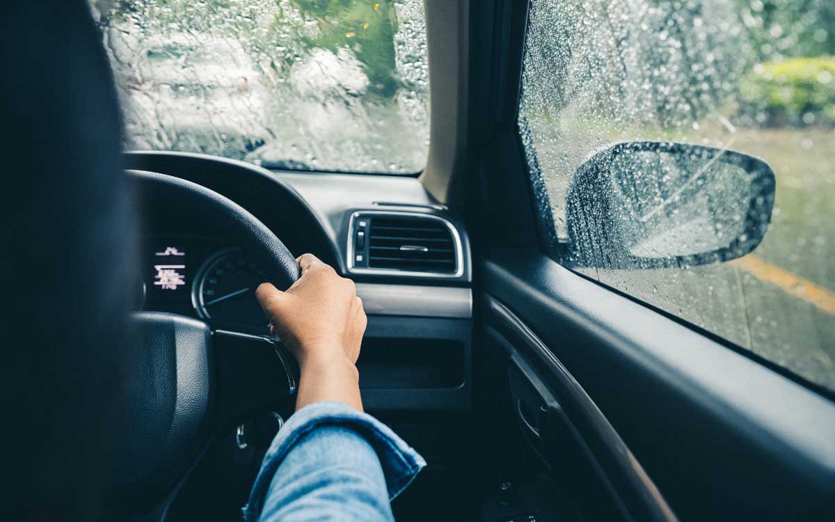 The perspective of a driver in a vehicle during a rainstorm, with hands on the steering wheel. The windshield is covered with raindrops, and the view outside is blurred, indicating poor visibility.