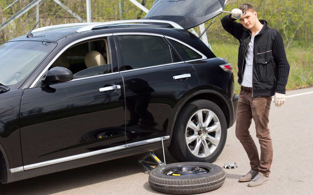 An adult male appears frustrated while standing next to a black SUV with a flat tire and a spare tire lying on the ground. The car's trunk is open, and the man is holding a tire iron to his head as if scratching his head.